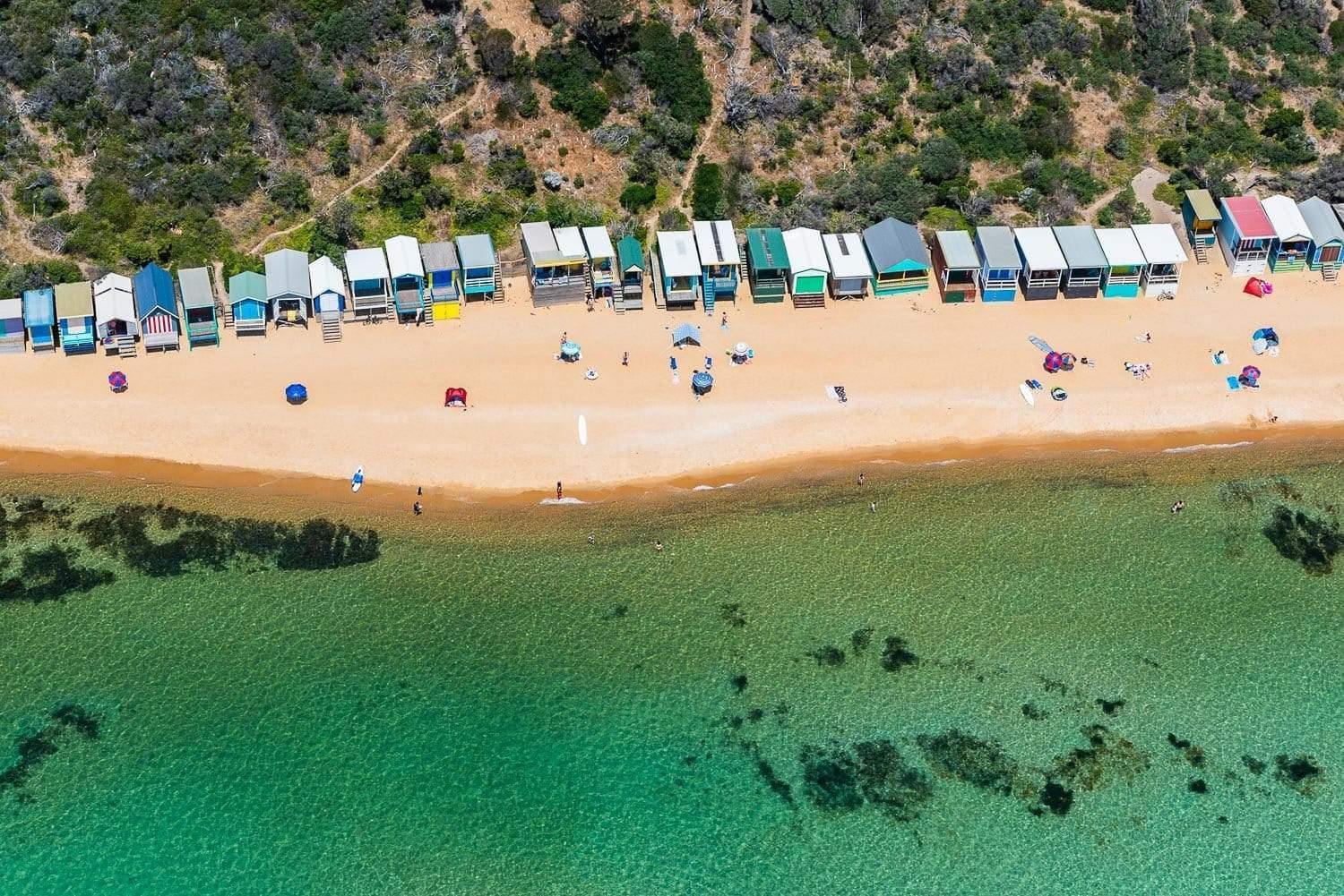 An aerial view of a green water beach with some colorful huts in a line, and some people and camps on the seashore with some plants and trees in the near background, Beachboxes Mt Martha Mornington Peninsula Victoria