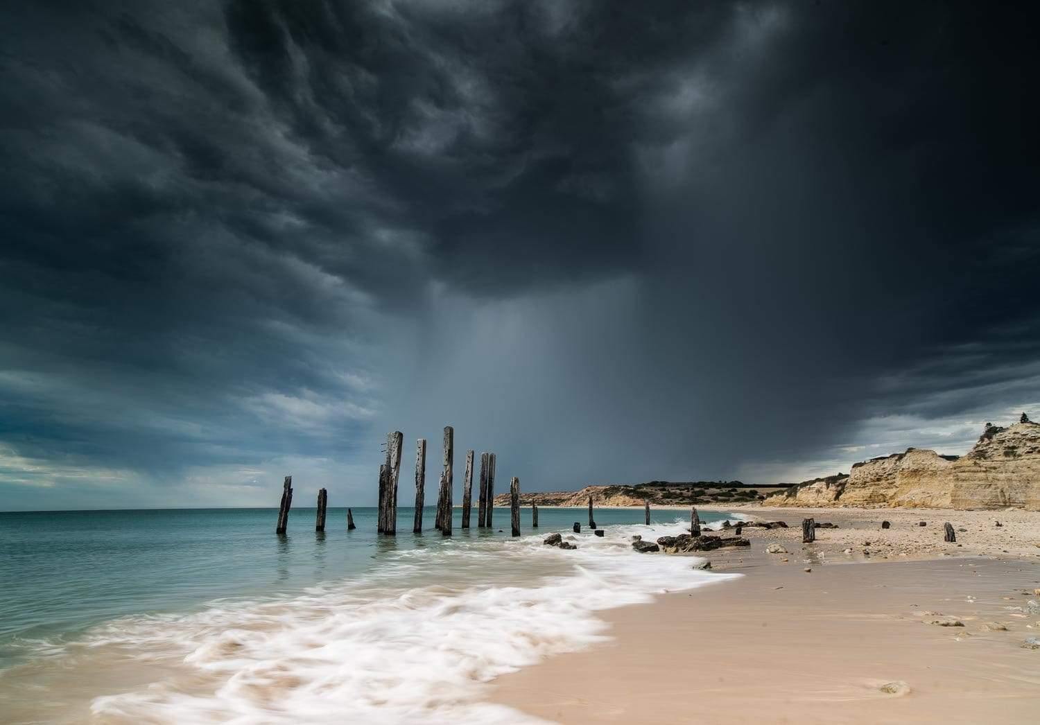 A sequence of long woody pillars standing in the water on the seashore with dark black clouds over them and some rocks in the right top corner, Blackened Skies Willunga SA Artwork