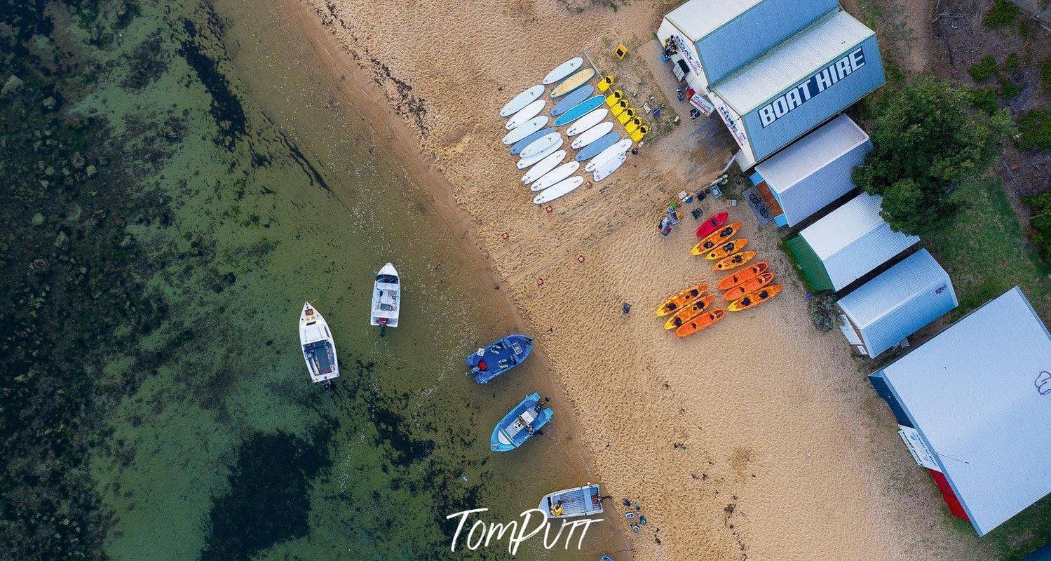 An aerial view of a seashore with some single-story houses and a lot of different sized boats standing on the land and some floating in the water,