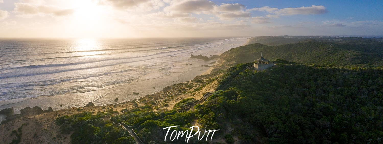 A long shot aerial view of a giant lush greenery hill point with a sea adjacent to it and a beautiful scene of the sun rising and spreading the sunlight, Coppins Lookout, Sorrento - Mornington Peninsula Victoria