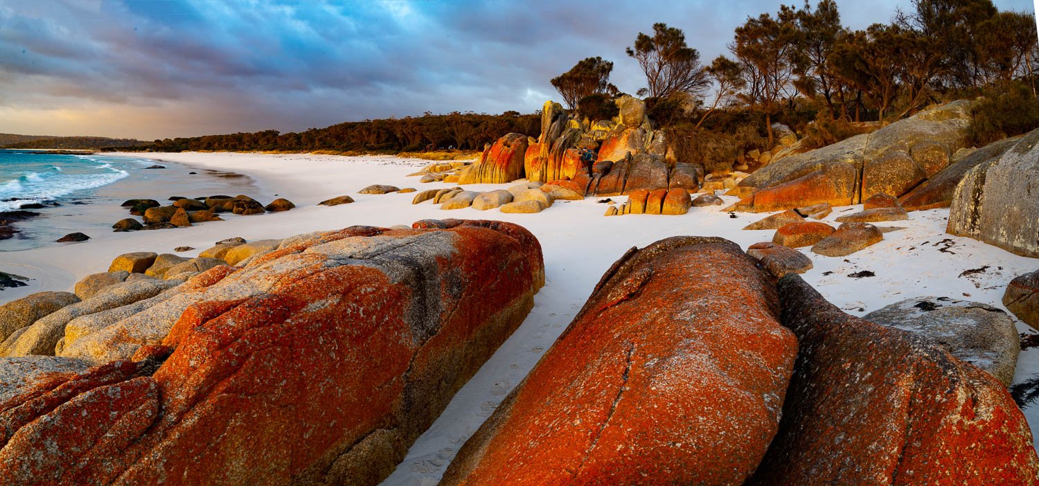 A beautiful landscape view of a seashore with white powder-like sand and some groups of large random shaped stones with red-colored texture and a group of trees in the background, Cosy Corner Sunshine, Bay of Fires