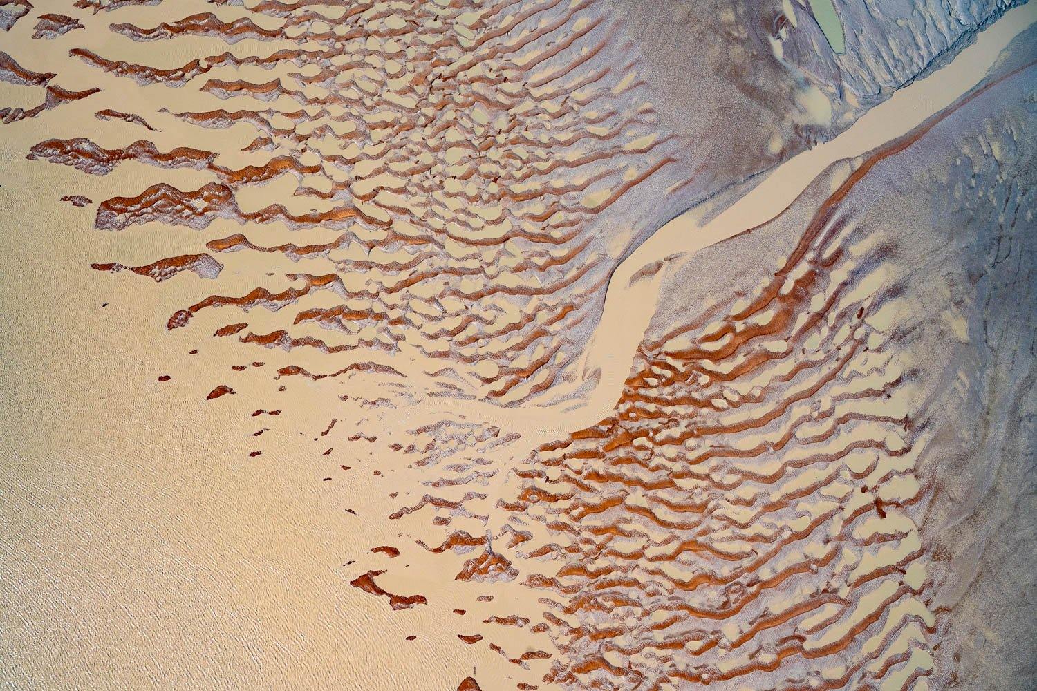 An aerial view of a desert-like land with a sequence of countless rounded zig-zag marks of brown color on it, and a thick pathway of sand color between the image connecting with the water, Derby #15 