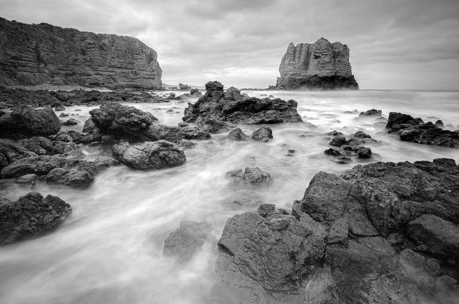 A black and white view of a sea corner with random shaped stones and a giant mountain wall with standing giant stone in the far background, Eagle Rock - Great Ocean Road VIC