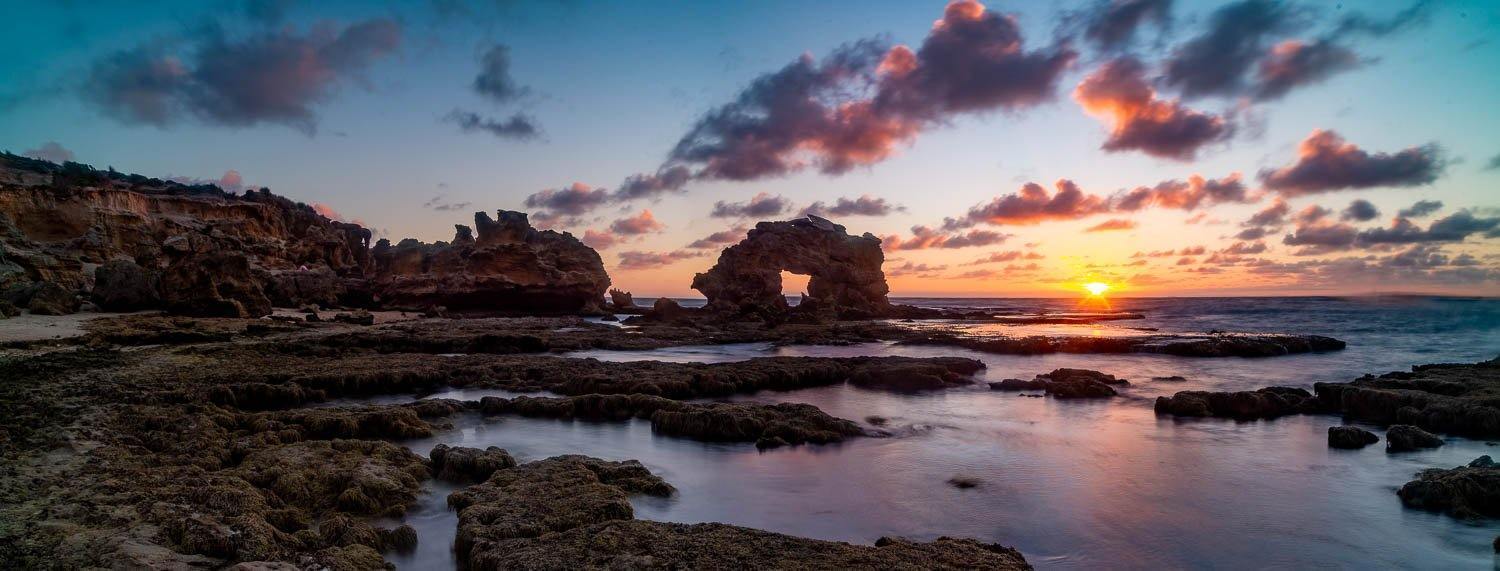 A wide sea shore with mountain stones, a large standing hollow mountain rock in the background, Keyhole Rock Sunset, Blairgowrie - Mornington Peninsula VIC 