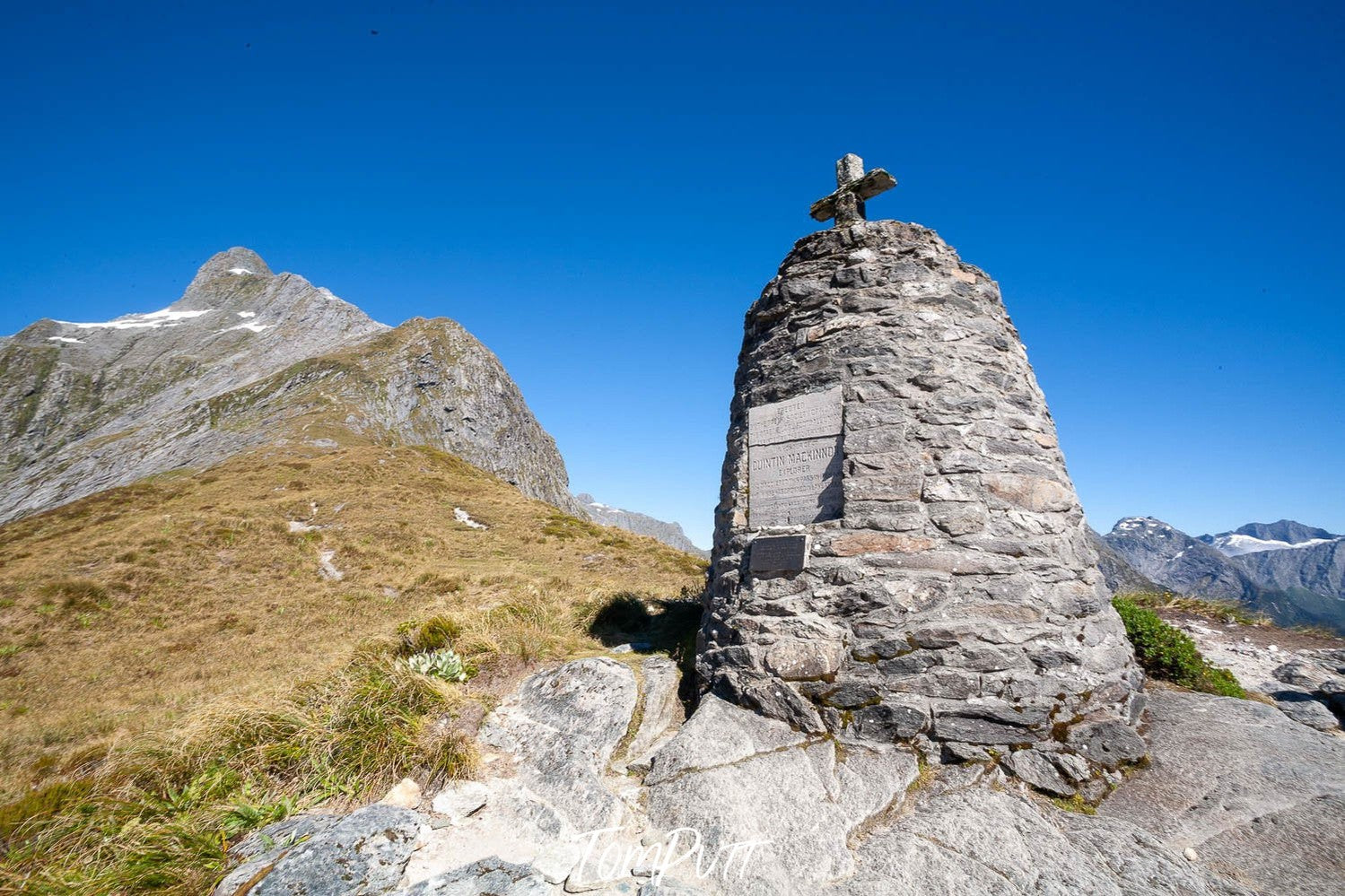 A standing stone with a plus sign on it in a greeny hill area, MacKinnon Pass Monument, Milford Track - New Zealand