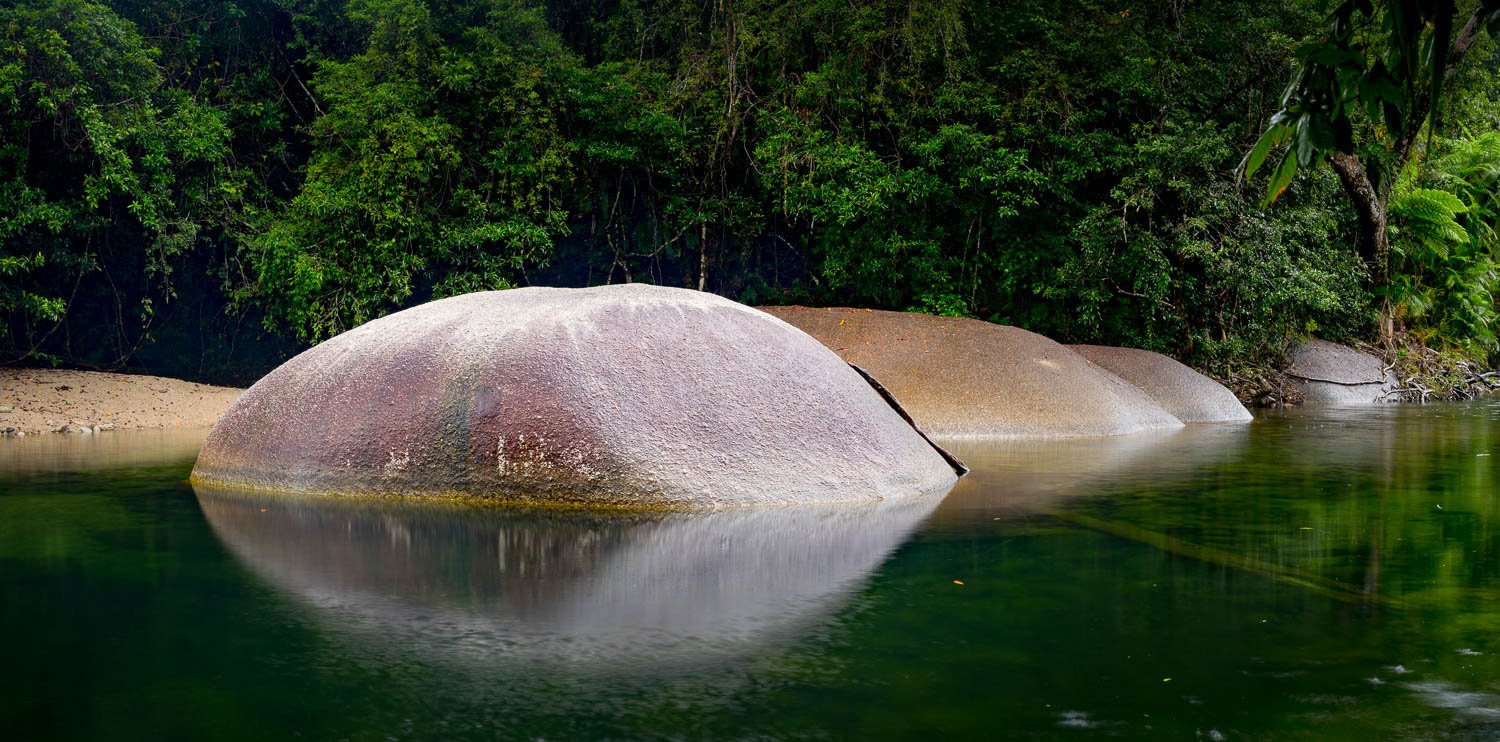 A series of round-shaped stones in the water creating clear half reflections in the water, with lush greenery behind them, Babinda Boulders Reflections, Far North Queensland