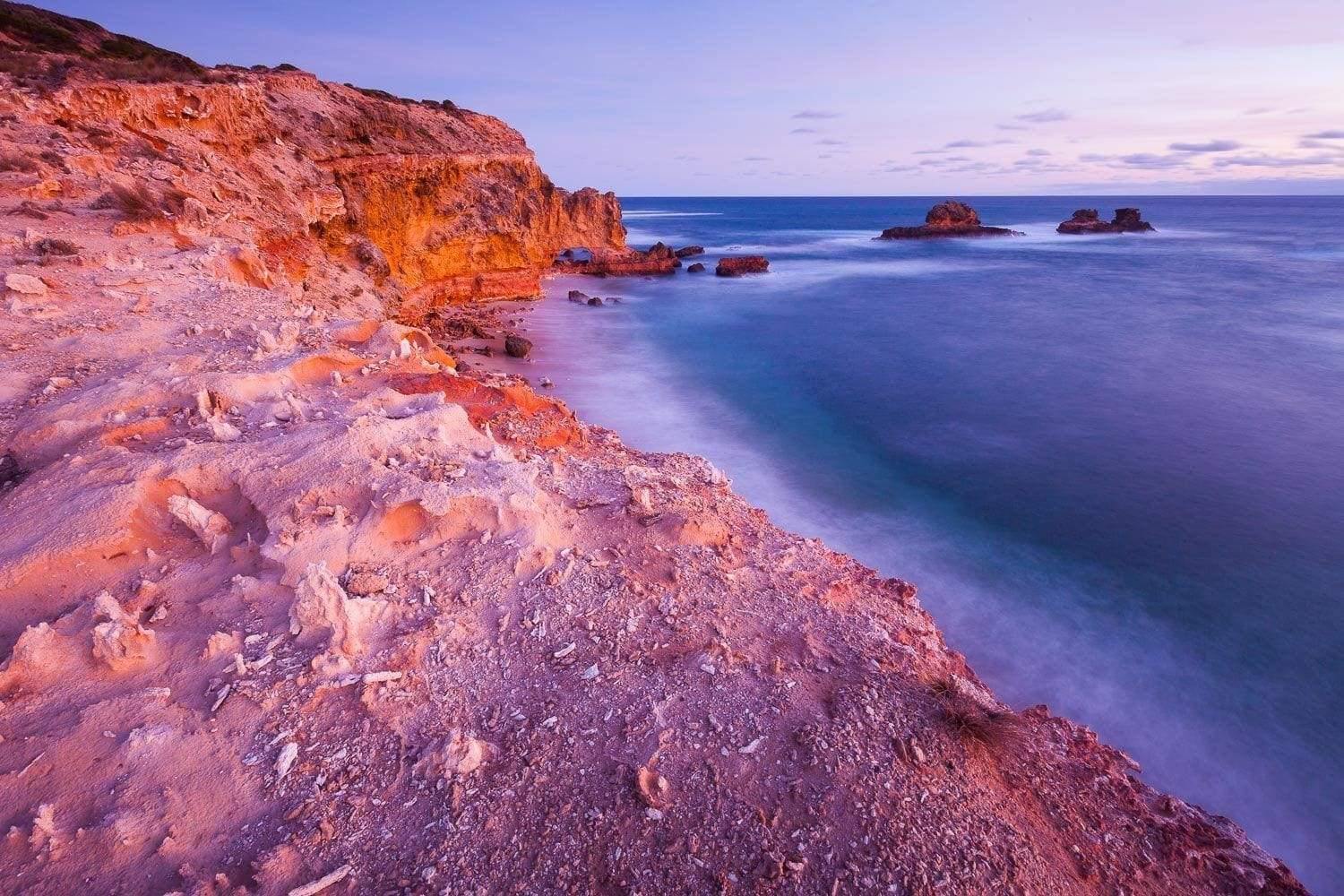 A morning view of a giant mountain wall with a seashore connecting to it, some big mounts are standing in the water in the far background, Bay of islands - Mornington Peninsula Victoria 