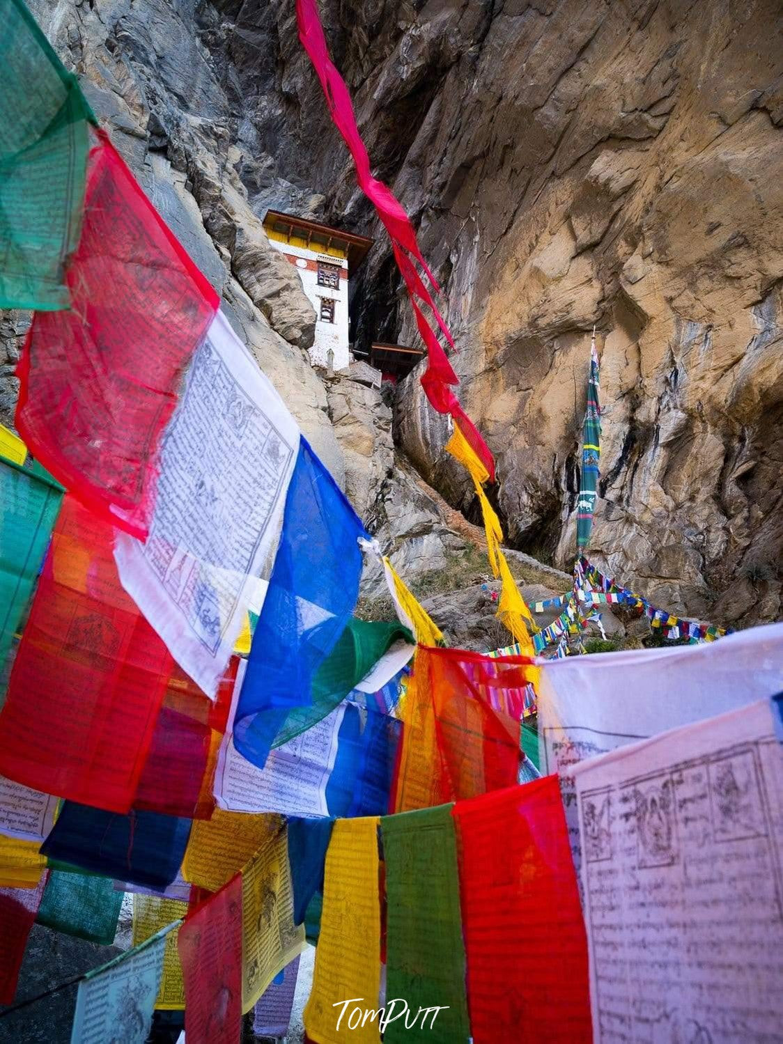 A close-up view of a series of colorful prayer flags and a prayer place on the rock in the near background, Bhutan Temple and Prayer Flags