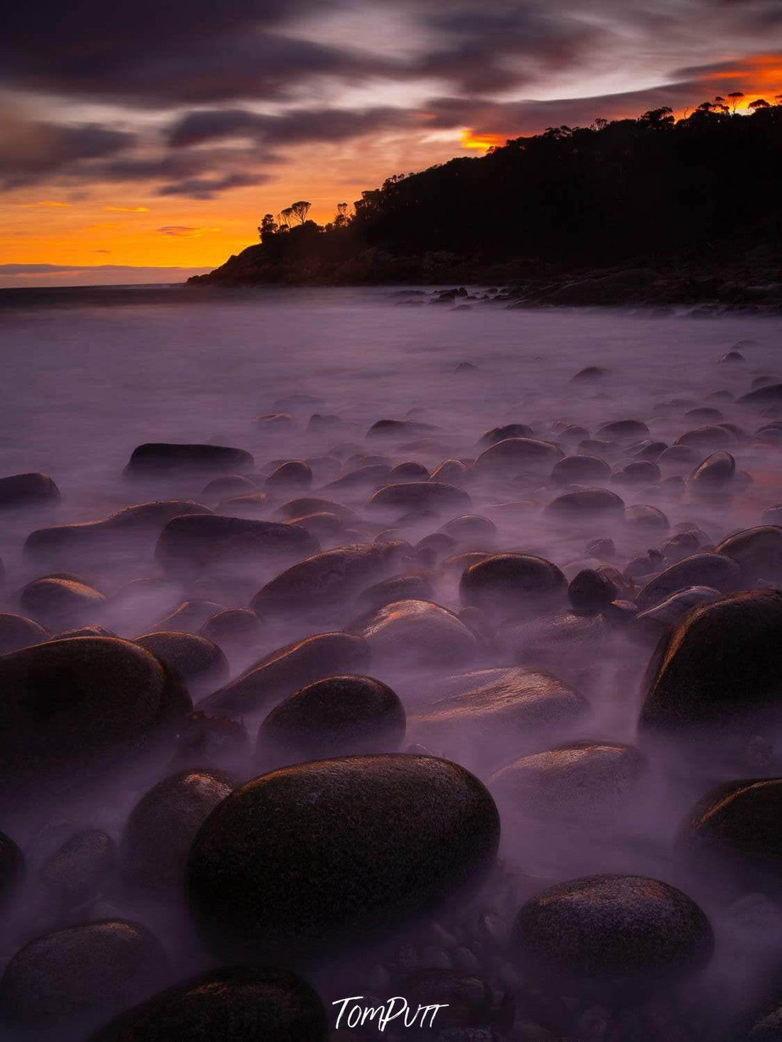 A group of lubricating rounded shape stones on the near-sea land, depicting like potatoes and the wet fog on them, a deep sunset view in the far background, Bluestone Bay sunrise, Freycinet