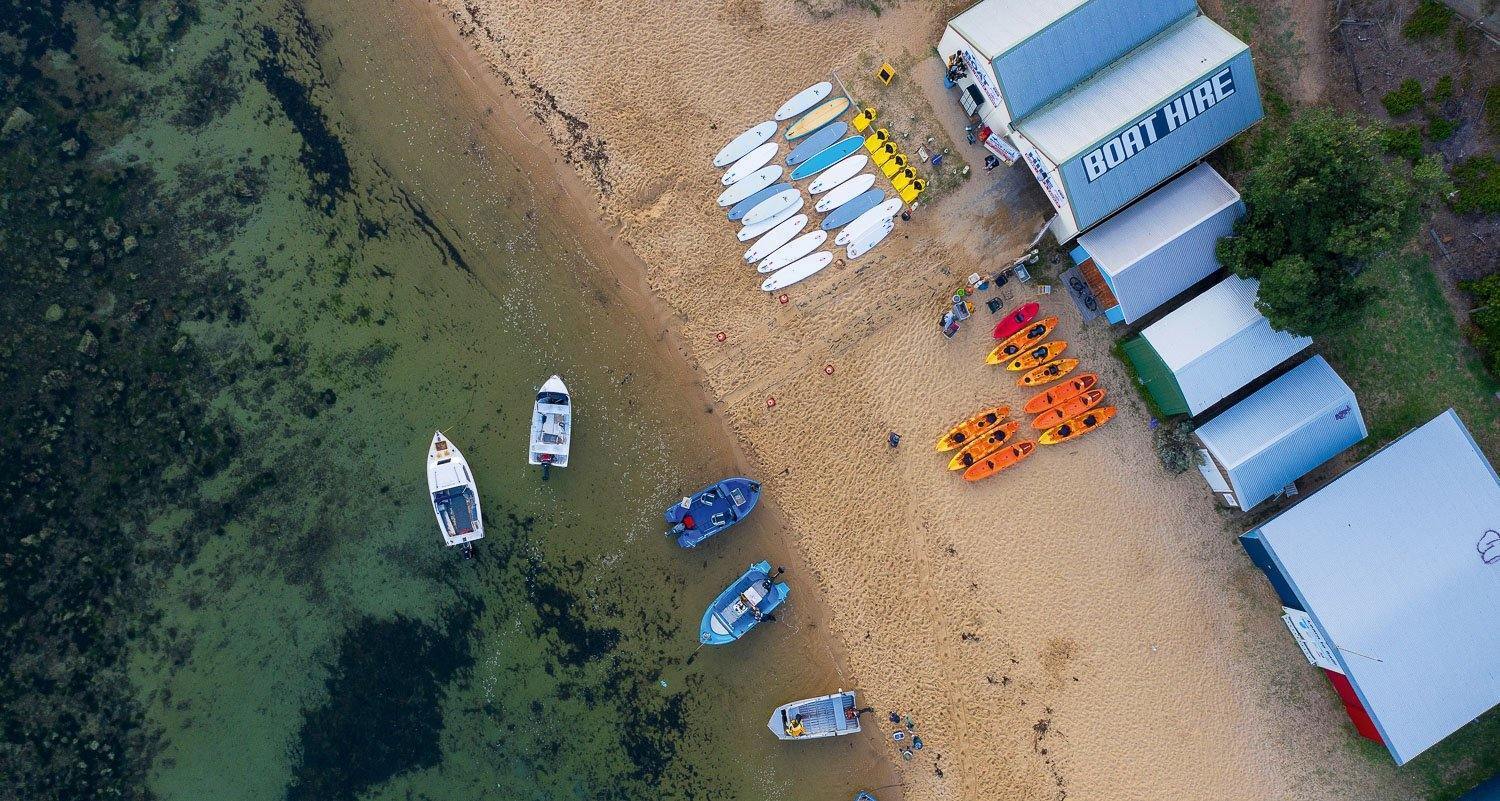 An aerial view of a seashore with some single-story houses and a lot of different sized boats standing on the land and some floating in the water, 