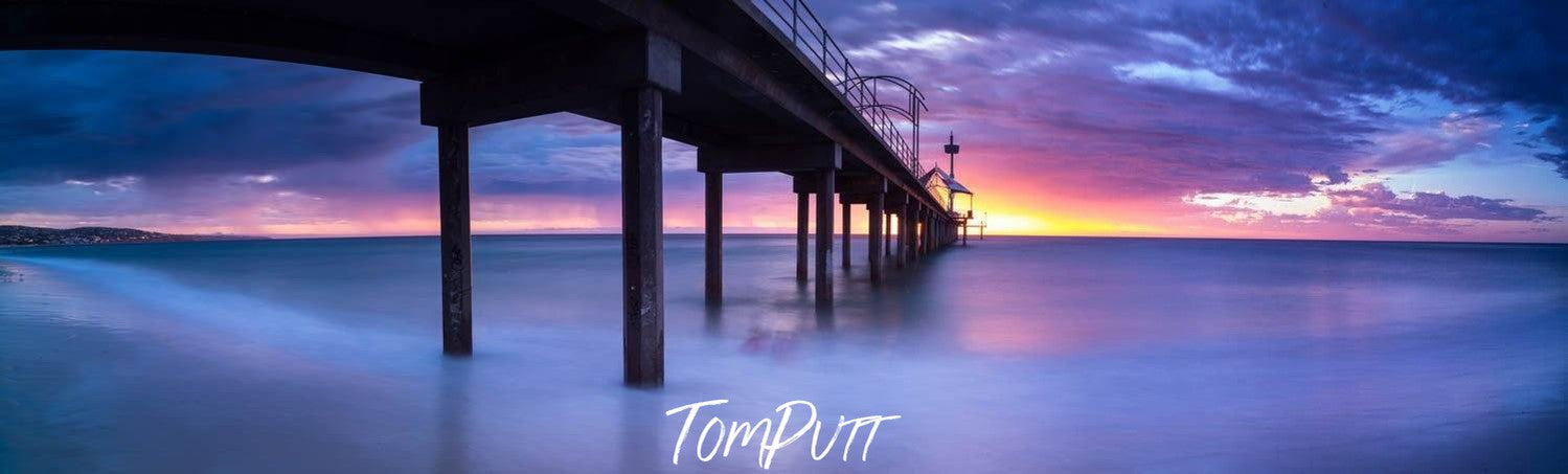 A dim light early morning view of an over-water bridge with its pillars in the water, and a purplish and blue shade on the sky with a little sunlight spreading in the far backgkround, Brighton Jetty - Adelaide SA