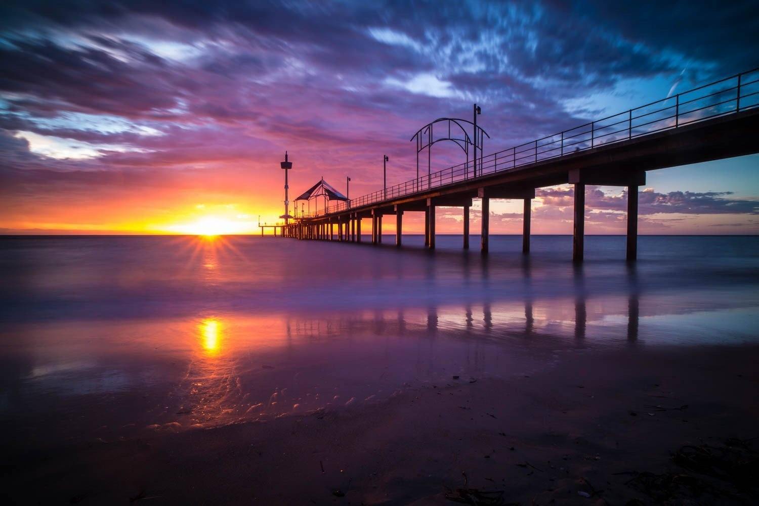 A long-shot view of an over-water bridge with its pillar in the water, some people waling on the bridge, and a tent is installed depicting a little cafe on the bridge, and a sunset effect forming a yellowish effect on the picture, Brighton Jetty Sunset - Adelaide SA