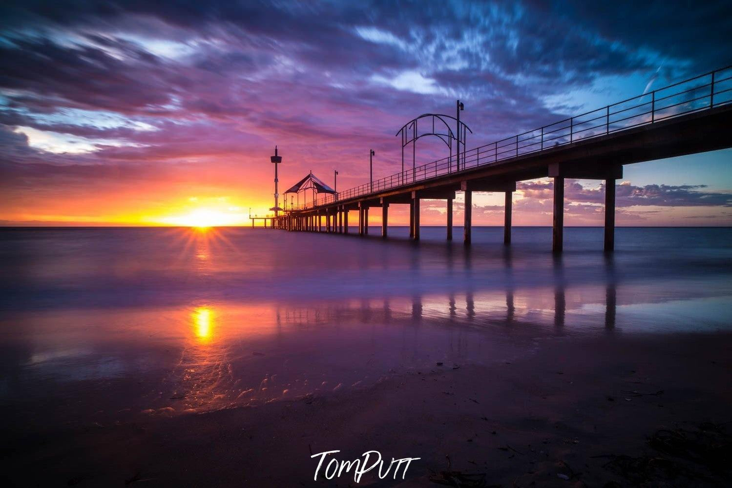 A long-shot view of an over-water bridge with its pillar in the water, some people waling on the bridge, and a tent is installed depicting a little cafe on the bridge, and a sunset effect forming a yellowish effect on the picture, Brighton Jetty Sunset - Adelaide SA