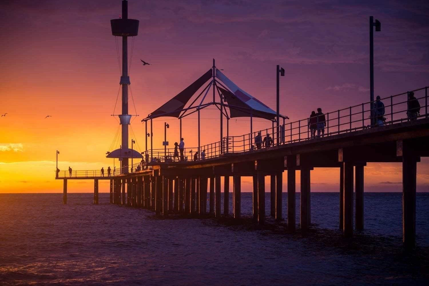 A beautiful view of an over-water bridge with its pillar in the water, some people waling on the bridge and a tent is installed depicting a little cafe on the bridge, Brighton Jetty Sunset - Adelaide SA