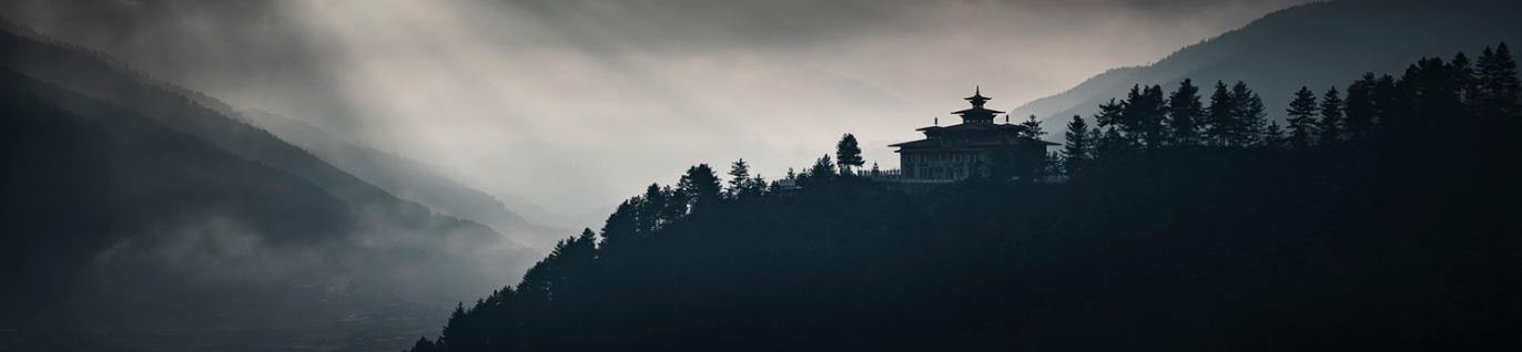 A dark view of a fort on the hillside of a mountain with some greenery, with some moonlight, and foggy effect, and a couple of giant blurred mountains in the far background, Bumthang Zhong - Bhutan