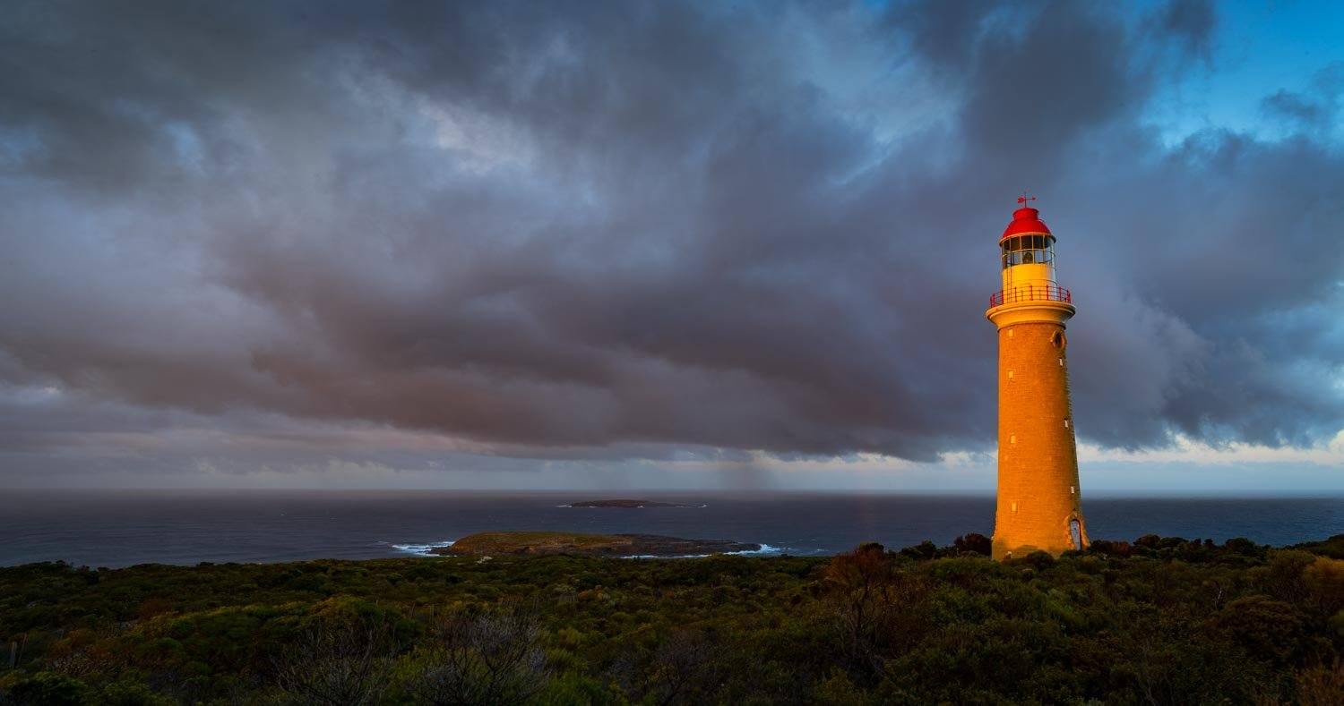 A beautifull lighthouse with hot shades with a long green field in the foreground, and a group of dark grey clouds over the scene, Cape Du Couedic Lighthouse - Kangaroo Island SA
