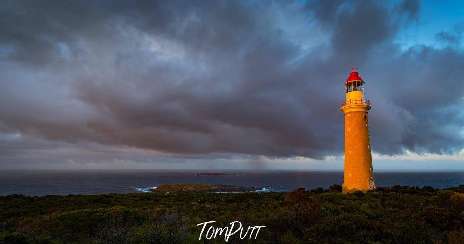 A beautifull lighthouse with hot shades with a long green field in the foreground, and a group of dark grey clouds over the scene, Cape Du Couedic Lighthouse - Kangaroo Island SA