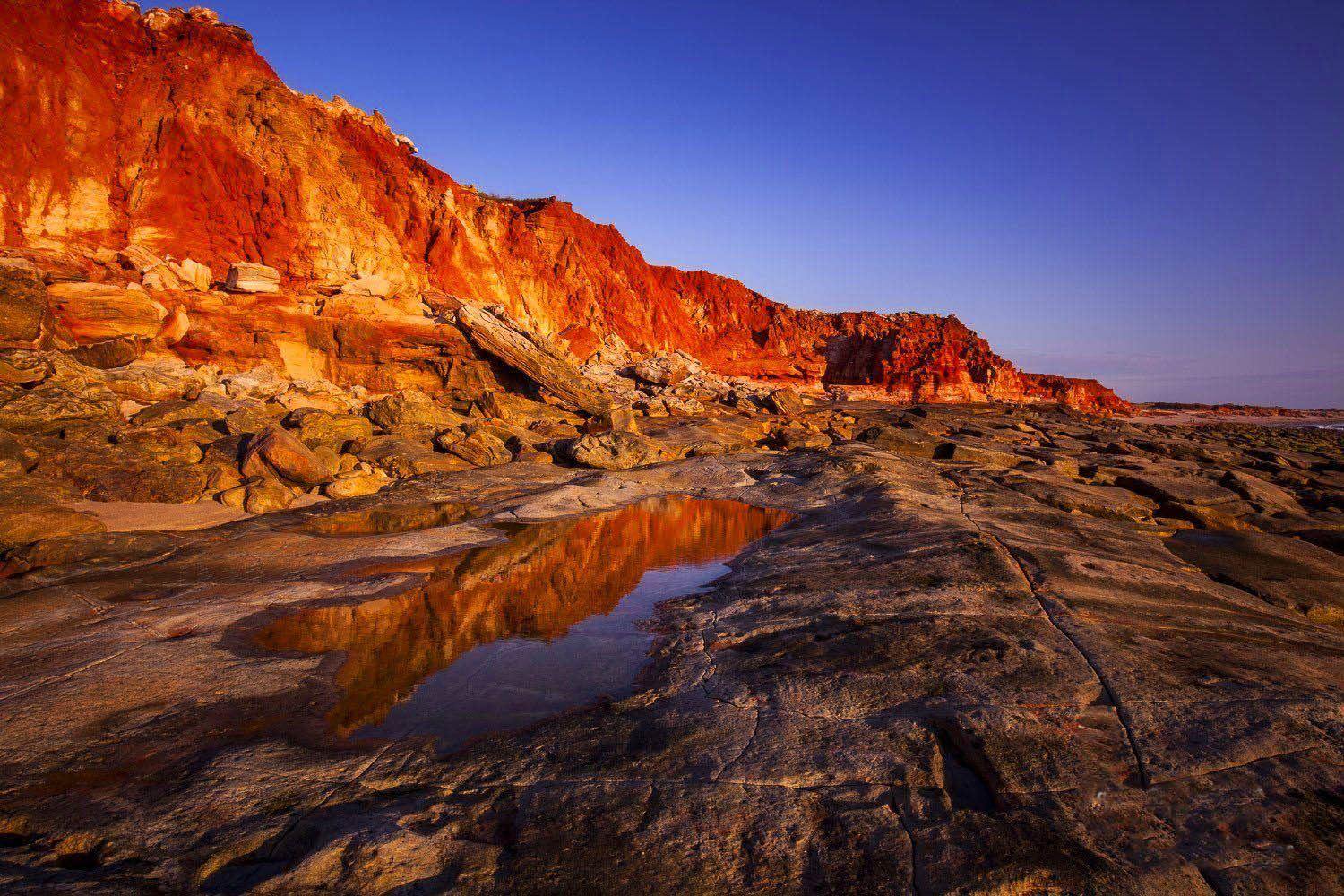 A long view of a long mountain wall with the shades of shiny red and orange, depicting a burning fire phenonema, a little water on the land with a clear reflection of the burning mountain wall, Cape Leveque - The Kimberley