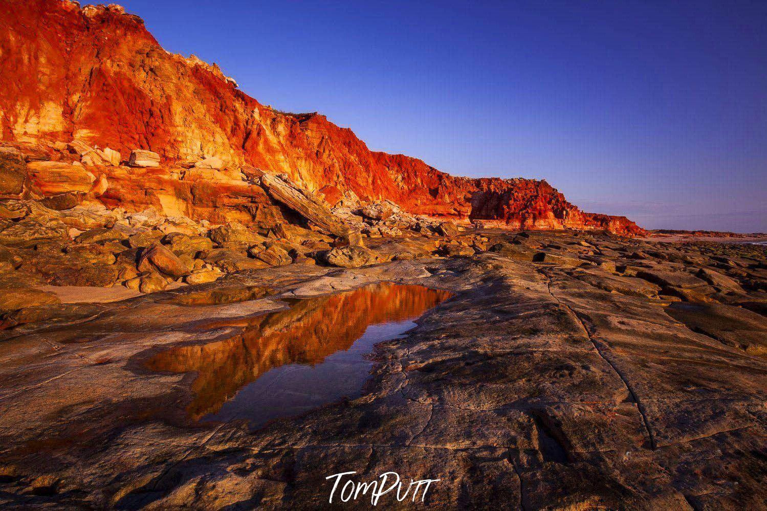 A long view of a long mountain wall with the shades of shiny red and orange, depicting a burning fire phenonema, a little water on the land with a clear reflection of the burning mountain wall, Cape Leveque - The Kimberley