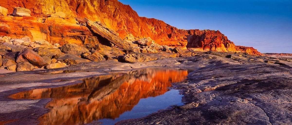 A long mountain wall with the shades of shiny red and orange, depicting a burning fire phenonema, a little water on the land with a clear reflection of the burning mountain wall, Cape Leveque - The Kimberley