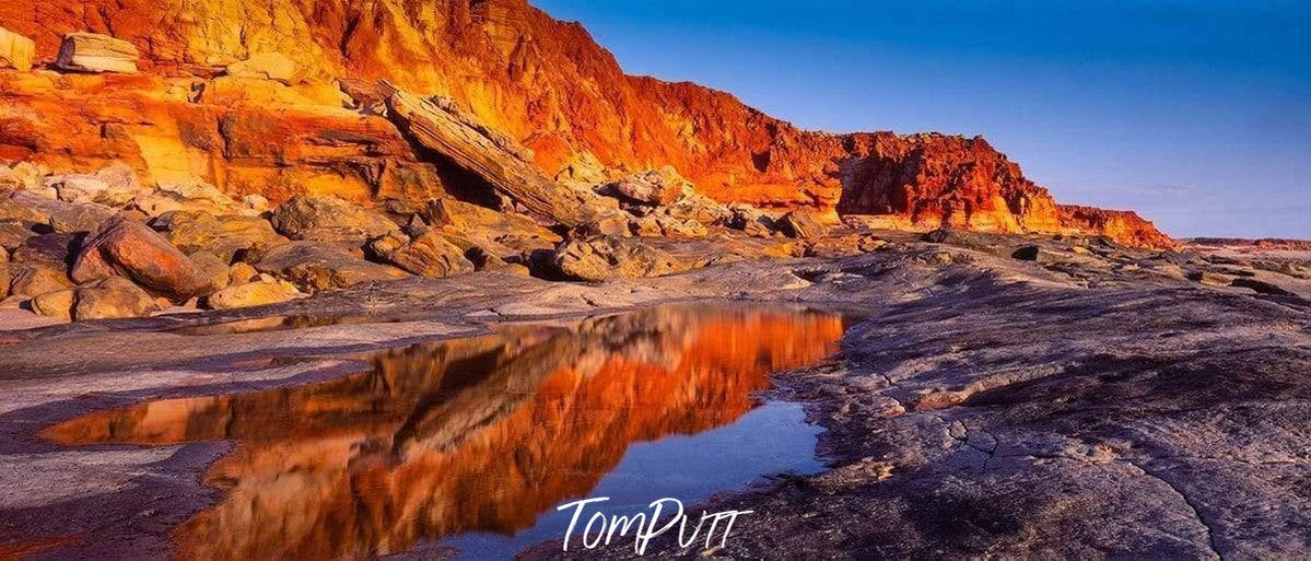 A long mountain wall with the shades of shiny red and orange, depicting a burning fire phenonema, a little water on the land with a clear reflection of the burning mountain wall, Cape Leveque - The Kimberley