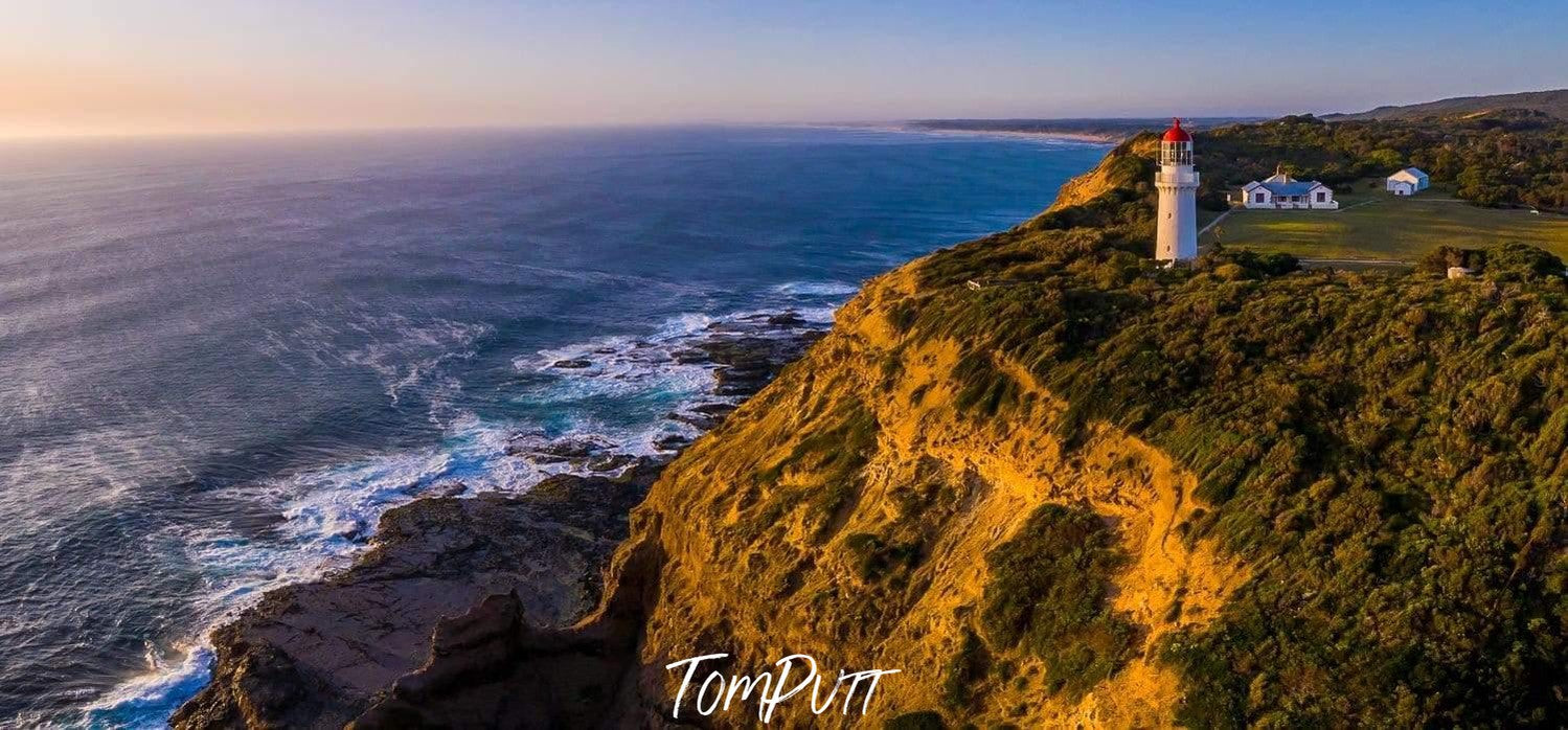 A long shot drone capture of the lighthouse on the corner of a grassy mound, and a seashore of an ocean below the grassy hill area, Cape Schanck Lighthouse - Mornington Peninsula