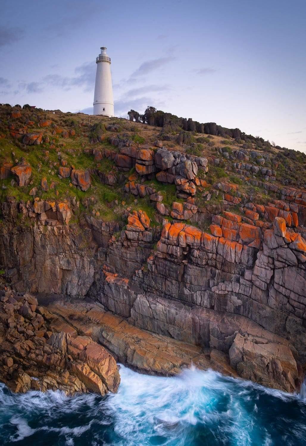 A portrait view of a beautiful lighthouse on the top of the green mound, and a cracky-shaped mountain wall in the foreground with bubbling water waves, Cape Willoughby Lighthouse - Kangaroo Island SA