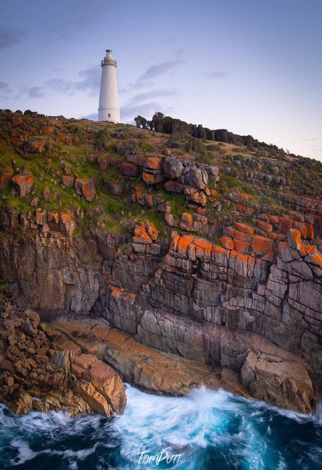 A portrait view of a beautiful lighthouse on the top of the green mound, and a cracky-shaped mountain wall in the foreground with bubbling water waves, Cape Willoughby Lighthouse - Kangaroo Island SA