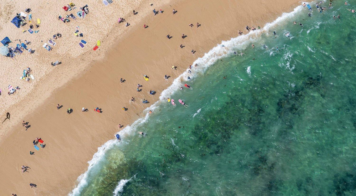 An aerial view of a deep sea-green colored beach connecting with the shore with so many huts and people on the beach enjoying their holiday, Catching Waves - Mornington Peninsula 