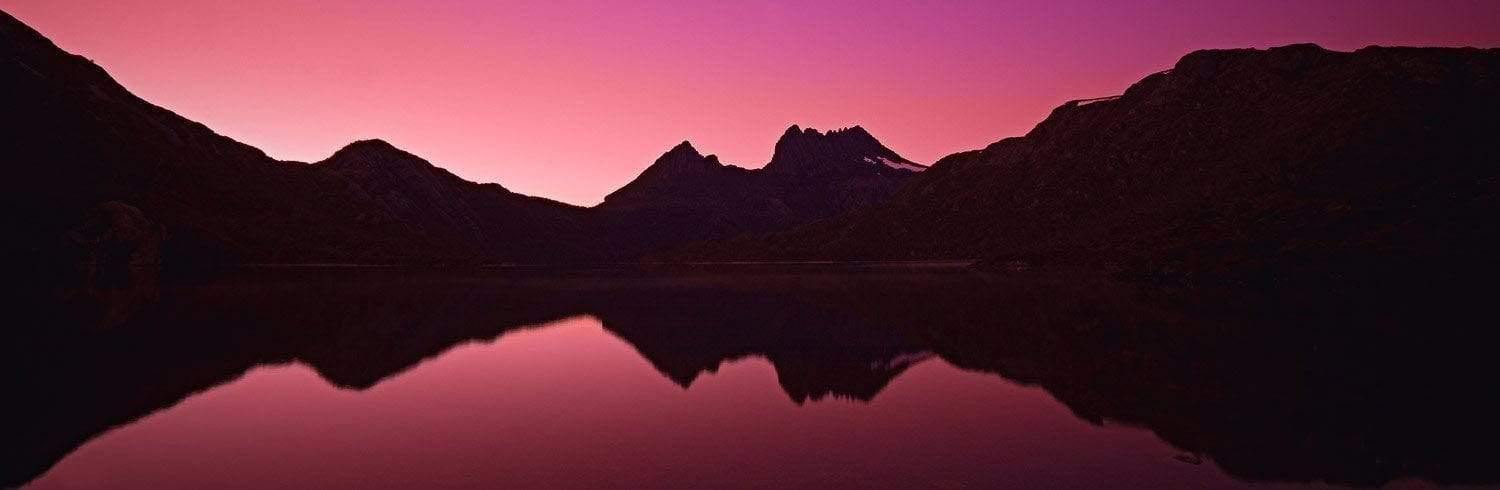 A dim light pinkish landscape view of a mountain-covered lake with a clear reflection of the mountains in the lake, Cradle Dawn - Cradle Mountain TAS