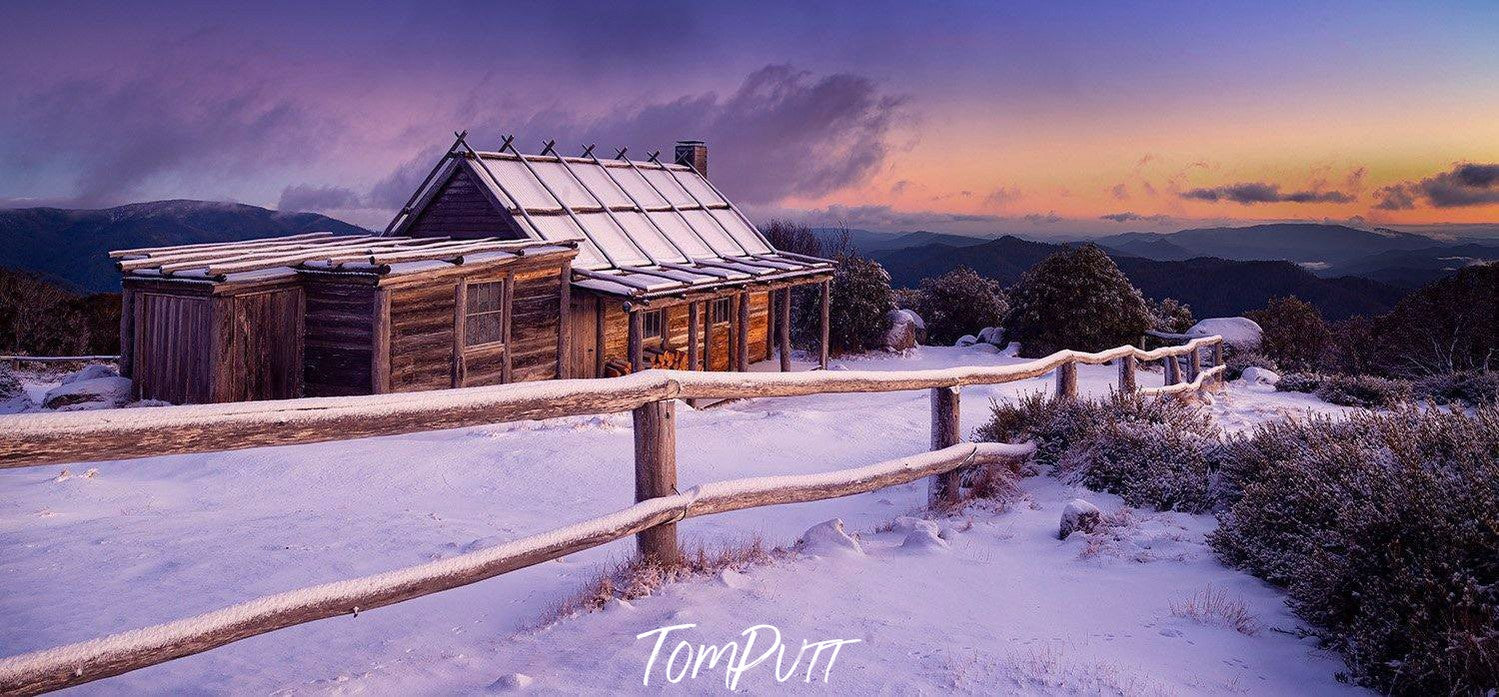 A snow area with a fence of wood and a small beautiful house with some dry bushes on the right corner, and a sunset view in the far background, Craig's Hut Dawn - Victorian High Country