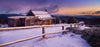 A snow area with a fence of wood and a small beautiful house with some dry bushes on the right corner, and a sunset view in the far background, Craig's Hut Dawn - Victorian High Country