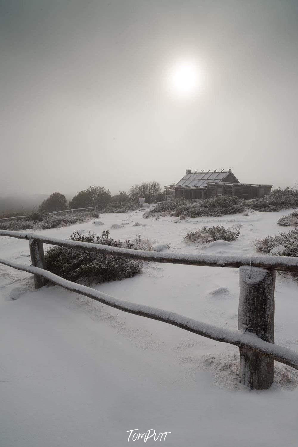 A beautiful snow-covered area with the fence of wood around it, some trees, and a small house inside, a shining sun with blurred sunlight on the scene, Craig's Hut Panorama - Victorian High Country