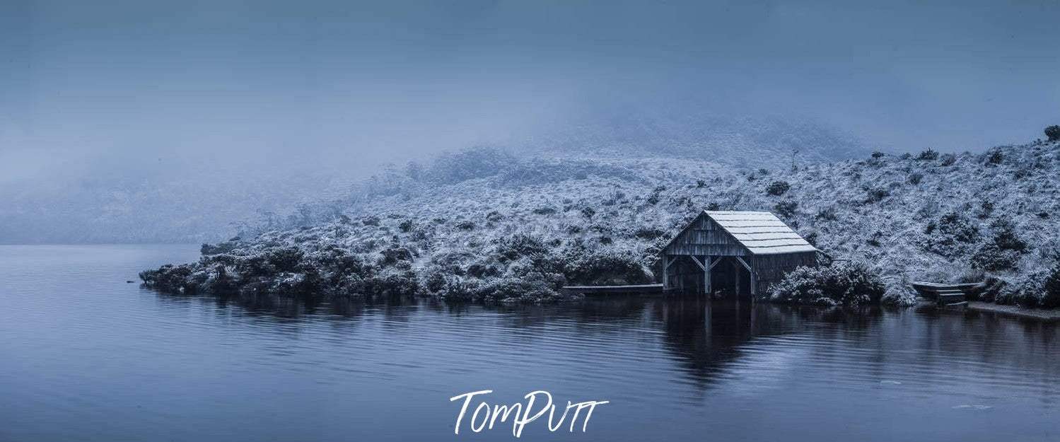 A small house with a greenery area covered with snow on the shore of a sea with a dim light effect of evening and fog, Boat Shed - Cradle Mountain TAS