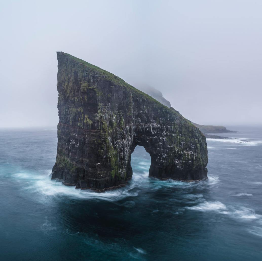 A giant stone shape in the sea with a hollow area similar to human legs, a steady flow of water with some dim light in the picture, Drangarnir, Faroe Islands