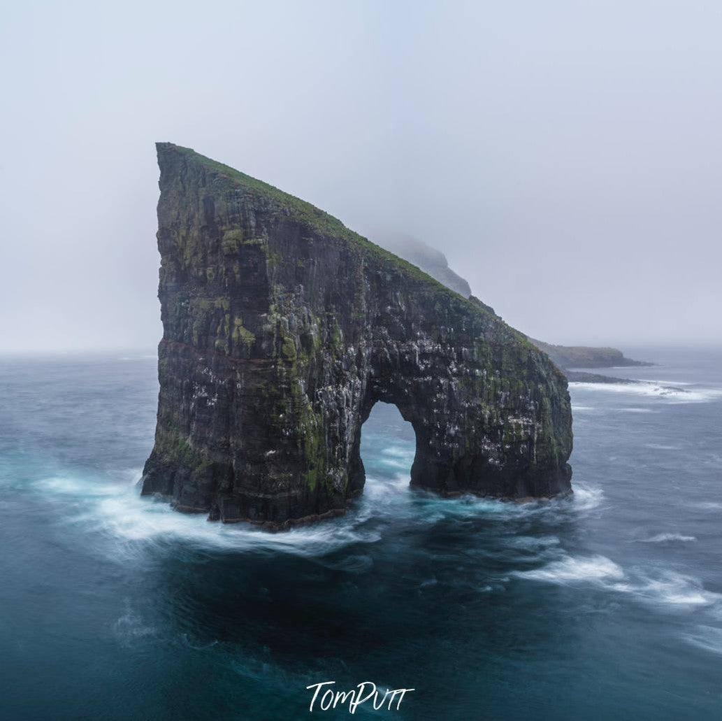 A giant stone shape in the sea with a hollow area similar to human legs, a steady flow of water with some dim light in the picture, Drangarnir, Faroe Islands