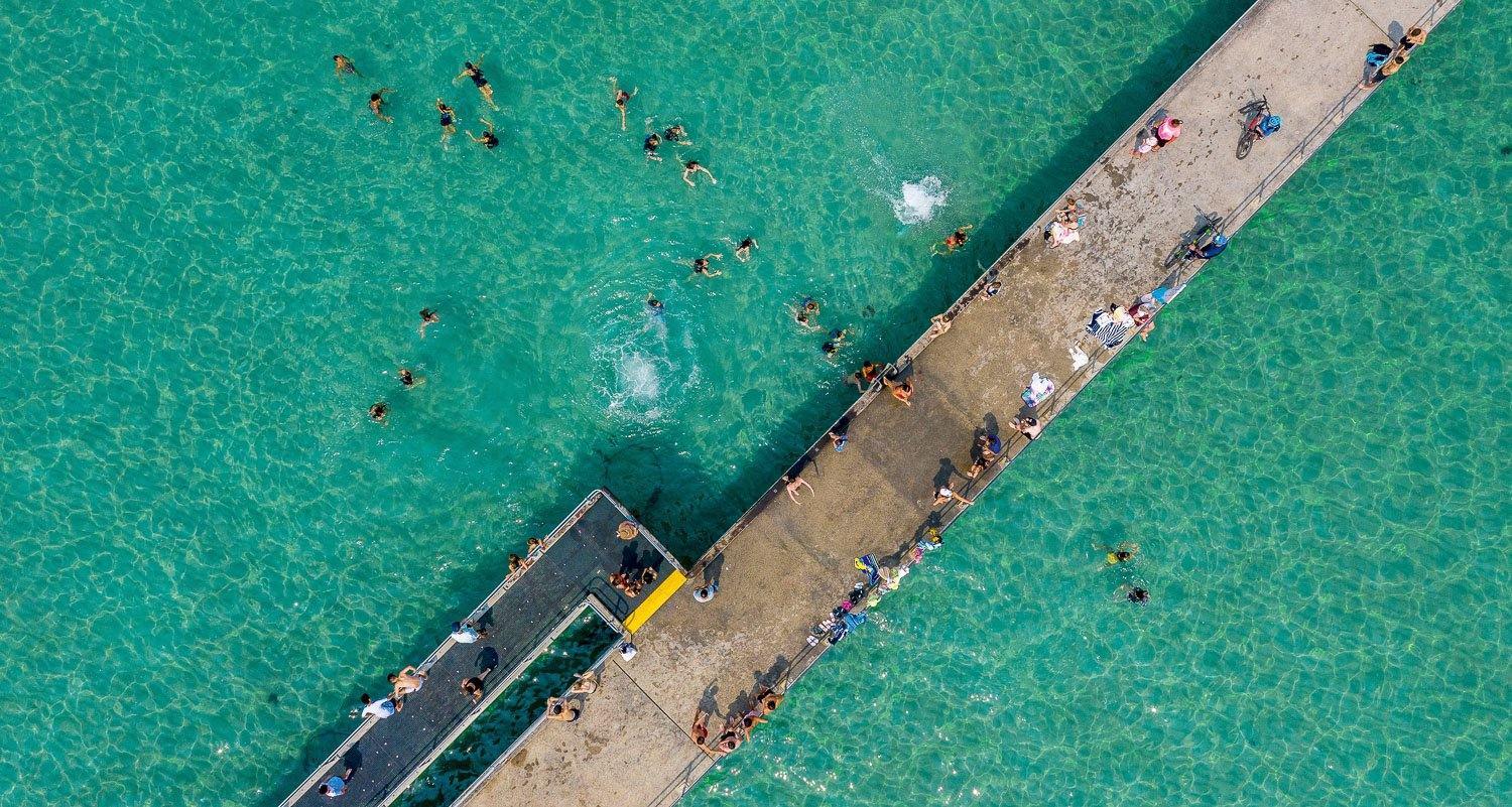 An aerial view of a bridge over the green-colored ocean, a number of people on the bridge and some on the water, Dromana Pier - Mornington Peninsula VIC