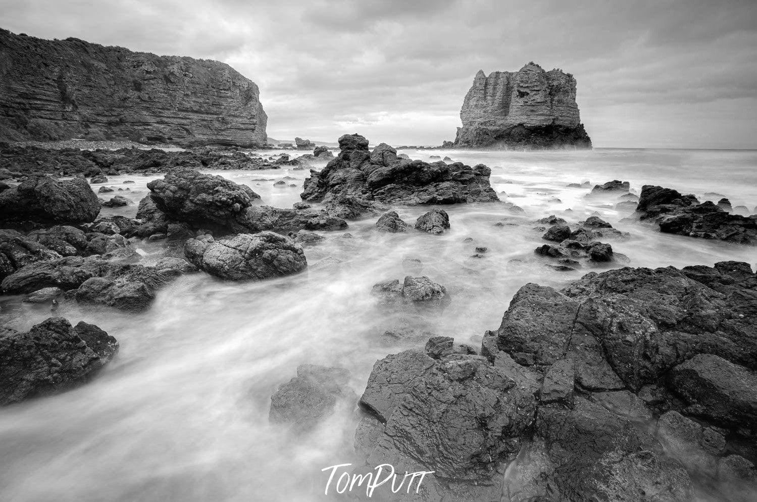 A black and white view of a sea corner with random shaped stones and a giant mountain wall with standing giant stone in the far background, Eagle Rock - Great Ocean Road VIC