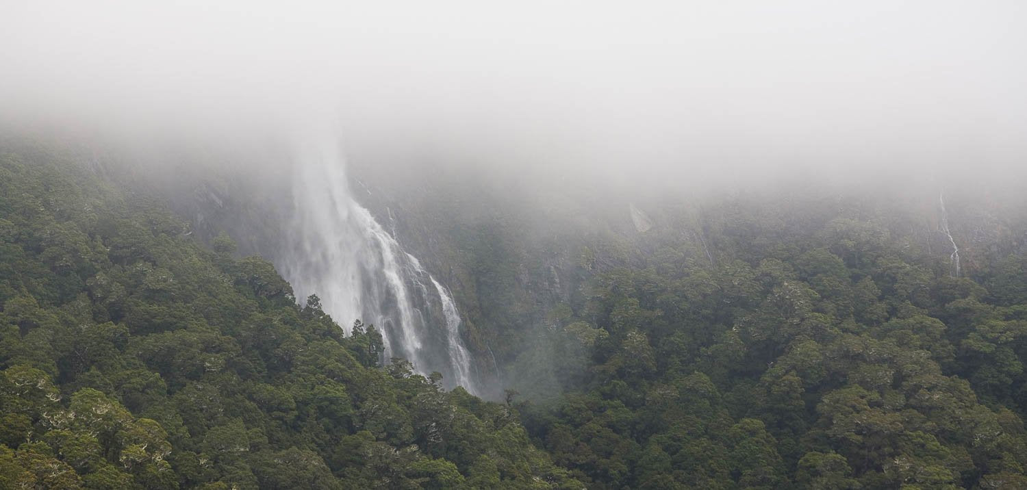 A hill area fully covered with greenery and a waterfall coming from the above, the top area is fully blurred due to a dense smoky effect, Earland Falls in flood, Routeburn Track - New Zealand