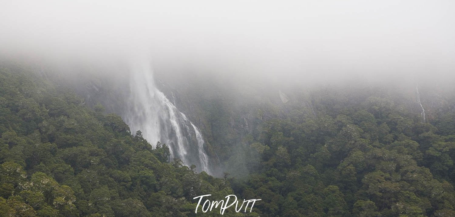 A hill area fully covered with greenery and a waterfall coming from the above, the top area is fully blurred due to a dense smoky effect, Earland Falls in flood, Routeburn Track - New Zealand