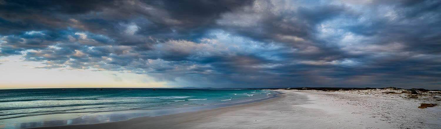 A landscape view of a beach with sky-blue water, Giant ice-blue and grey dense clouds over the scene, Eddystone Beach panorama, Bay of Fires