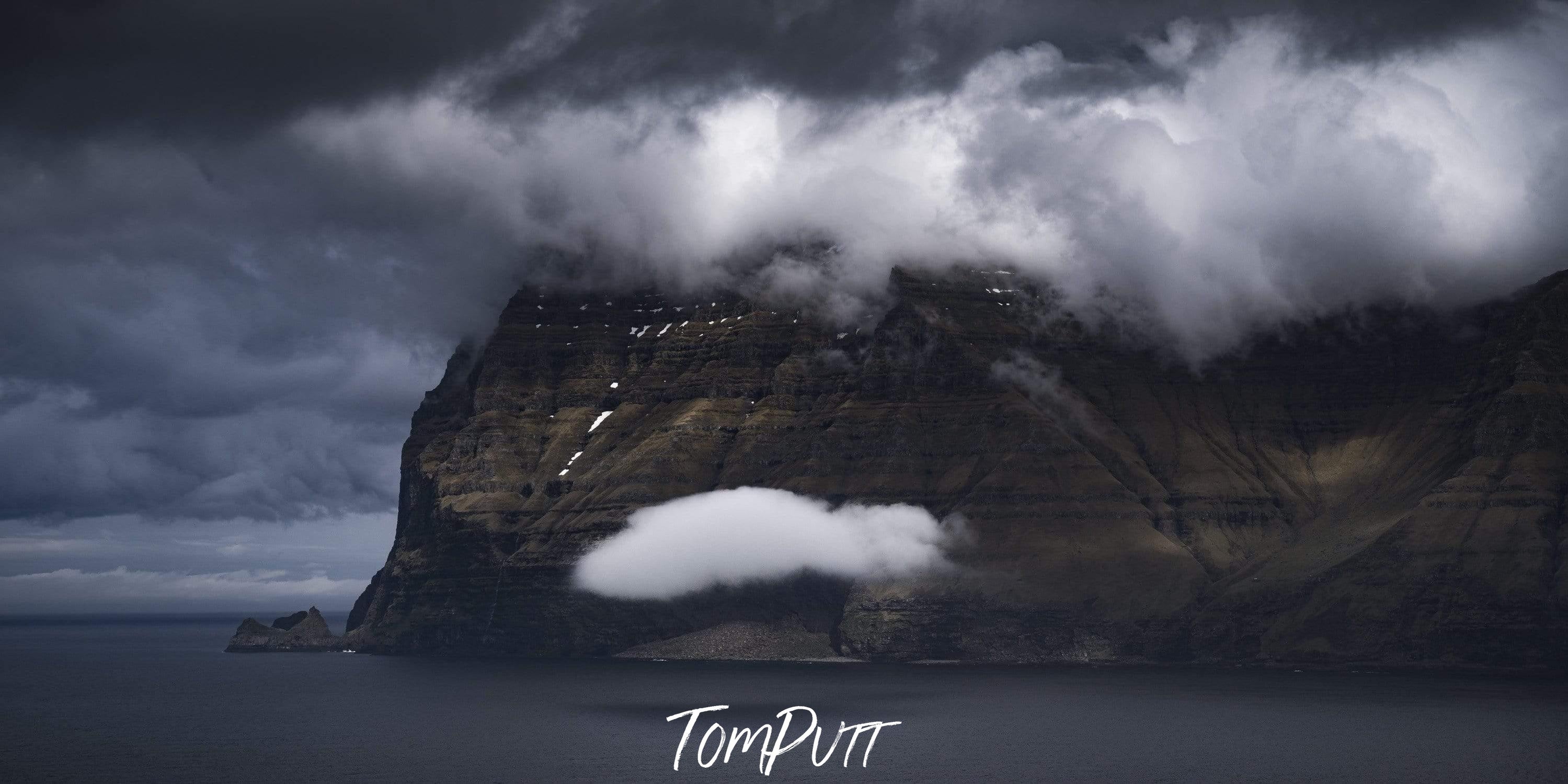 A giant mountain wall in the sea with a big hole in between, dense white clouds over it, Floating Along - Faroe Islands