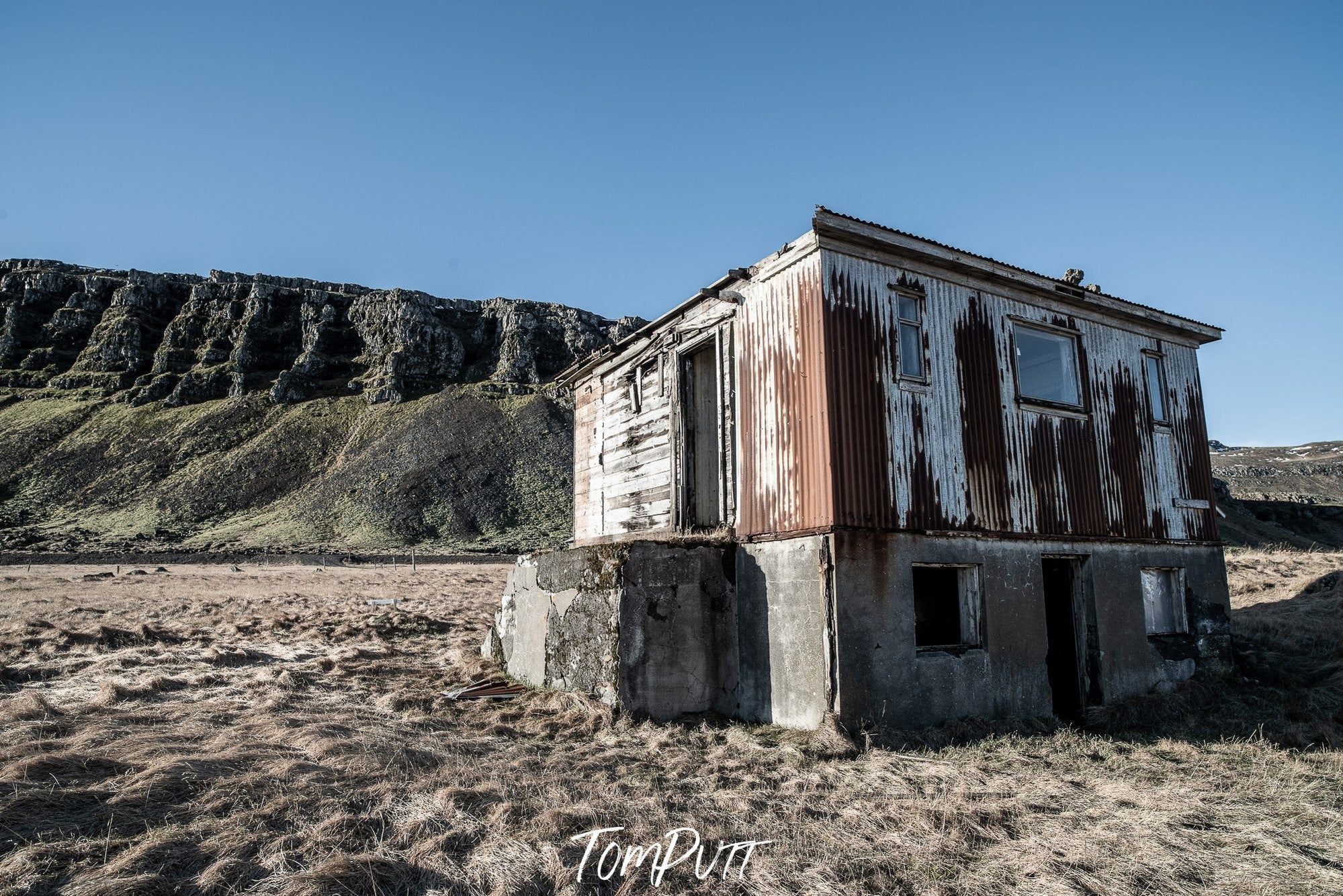 An old house on sand with long greeny mountains in the background, Iceland No.13