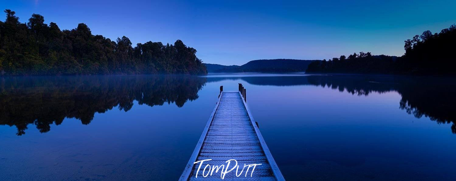 Dark view of a lake surrounded by greenery and a wooden bridge over the lake, and a clear reflection in the water, Lake Mapourika New Zealand Art