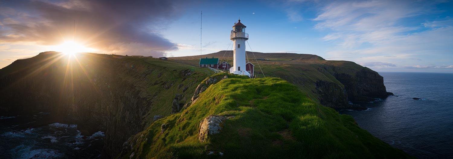 A long shot view of a lighthouse on a green hill point with an effect of sunrise in the far background, Lighthouse, Faroe Islands