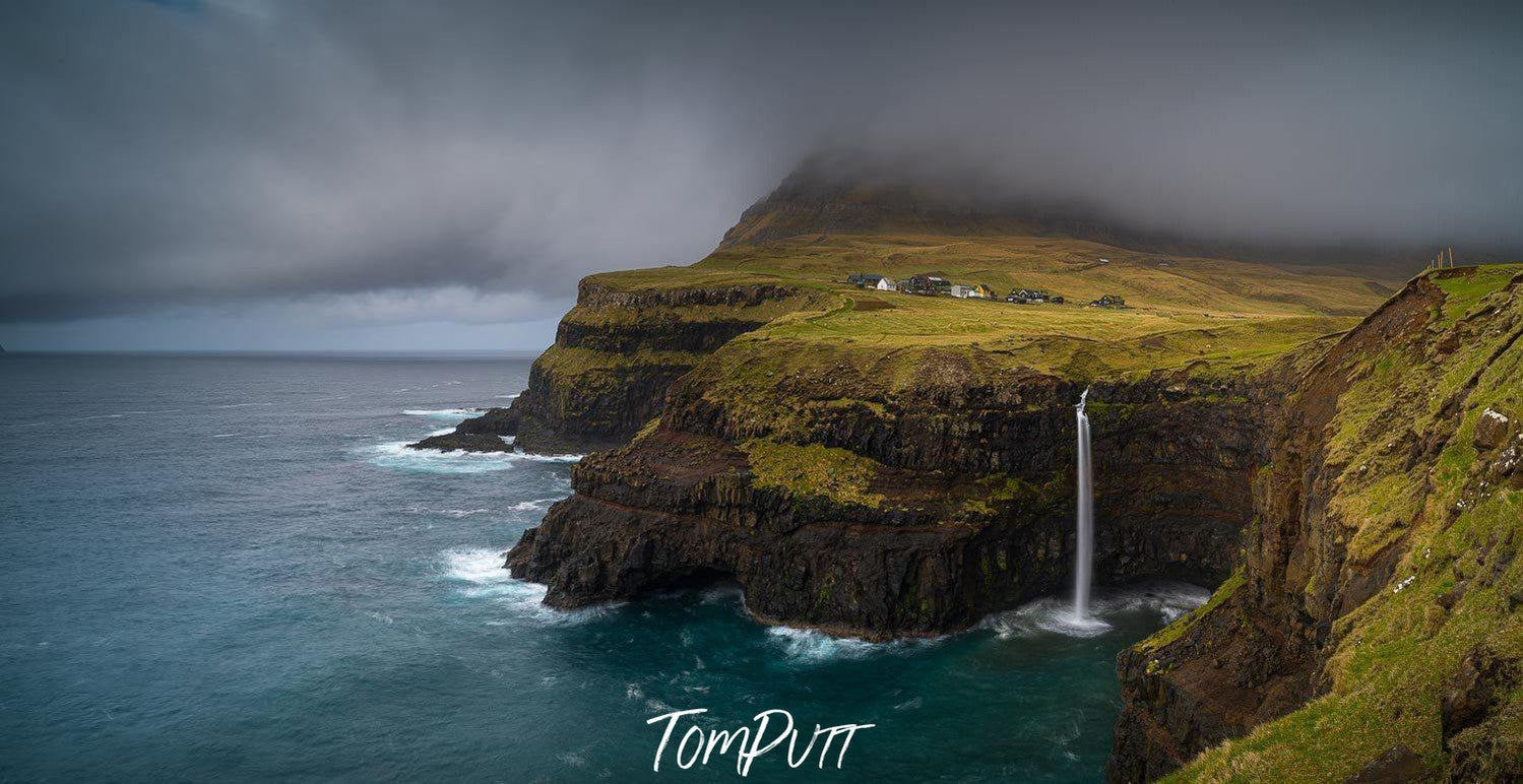 Long grassy mountains wall with heavy black clouds over and a lake corner below, Mulafossar Waterfall, Faroe Islands