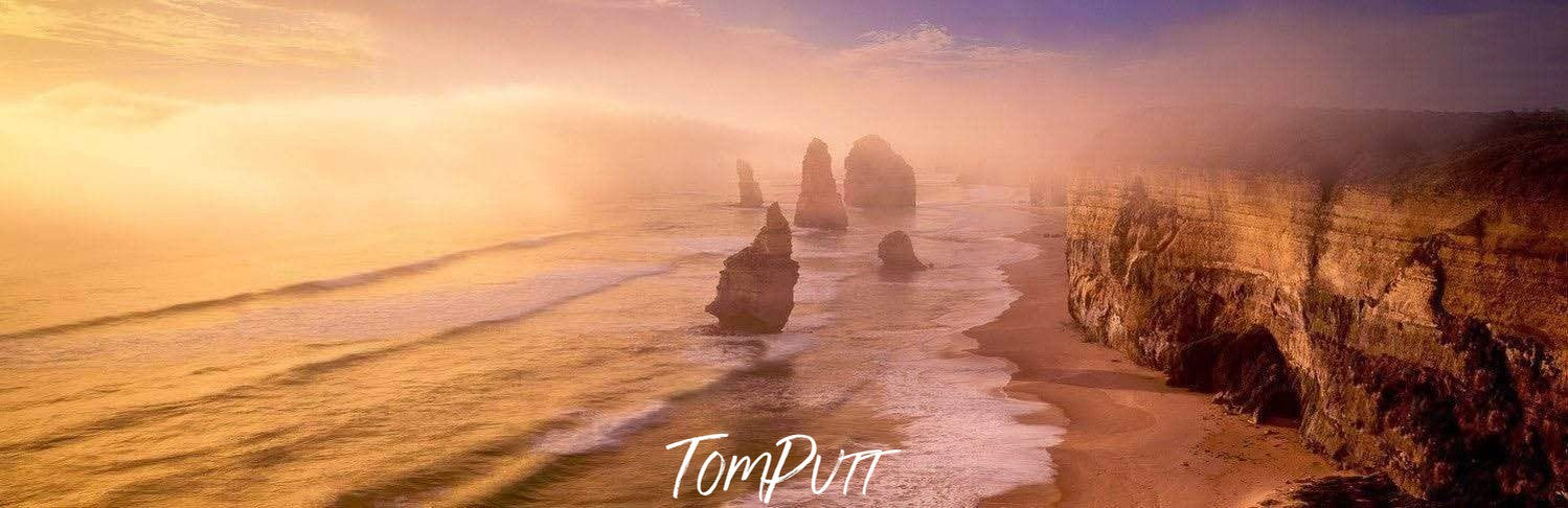 Long shot of a mountain wall and some standing stones below on the beach, Pillars in the Mist, 12 Apostles - Great Ocean Road VIC