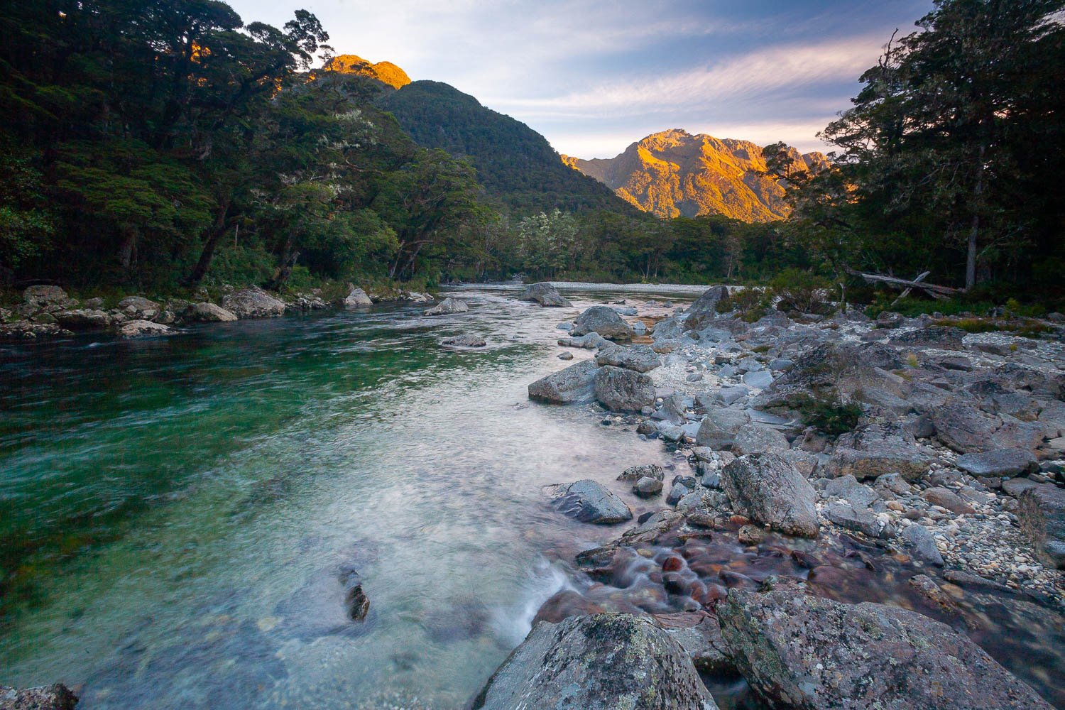 A watercourse with many stones aside, and a lot of greenery on both sides, and a shining mountain peak point in the background, The Clinton River and Earl Mountains at sunset, Milford Track - New Zealand