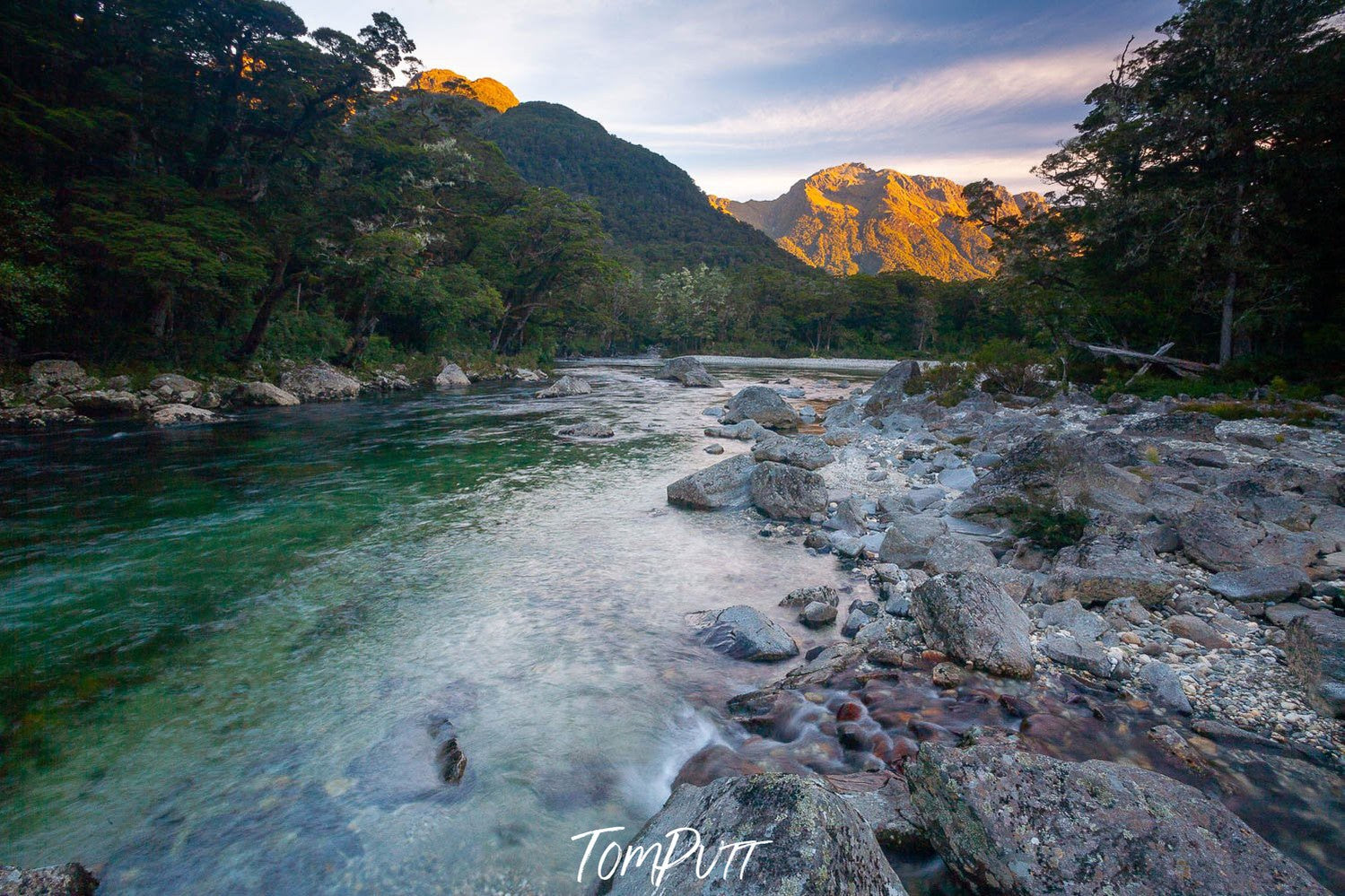 A watercourse with many stones aside, and a lot of greenery on both sides, and a shining mountain peak point in the background, The Clinton River and Earl Mountains at sunset, Milford Track - New Zealand