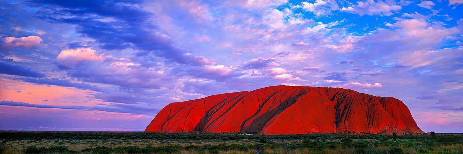 Long-shot of a ginat buring-orange mountain wall with purplish effect on the sky, Uluru Glow Red Centre NT
